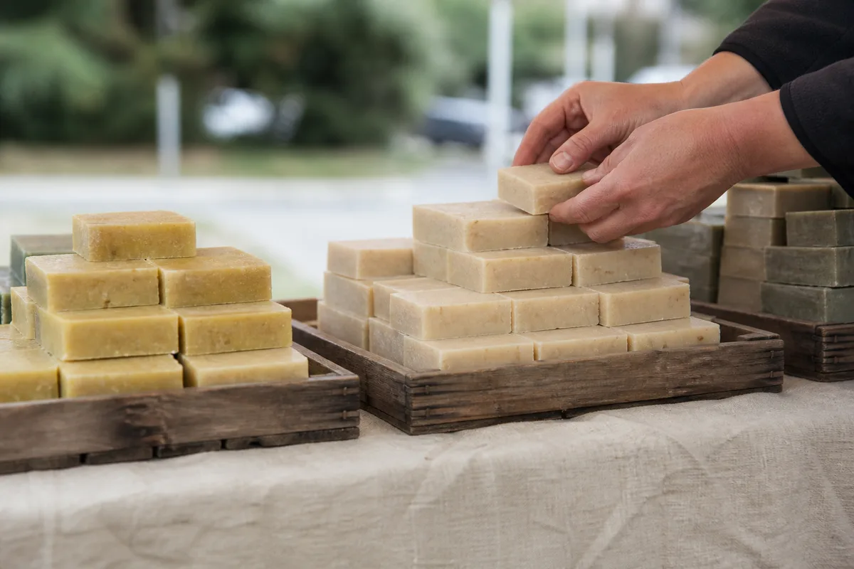 Étal de savons artisanaux empilés sur une nappe de lin, mains d&rsquo;une exposante replaçant une barre